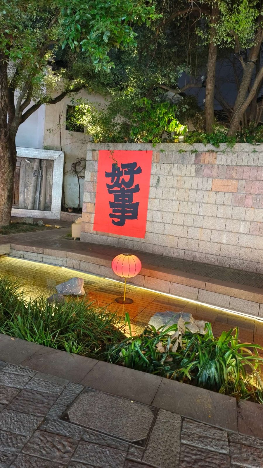 庭院石墙上贴着红底黑字的「好事」，水池边亮着一盏暖色纸灯笼